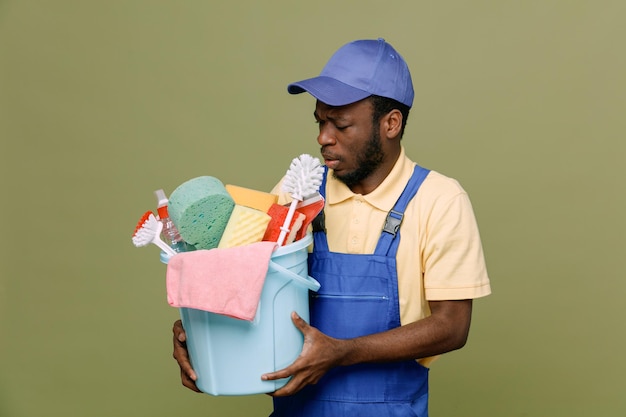 Seau de maintien tendu d'outils de nettoyage jeune homme nettoyeur afro-américain en uniforme avec des gants isolés sur fond vert
