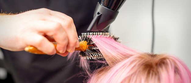 Séchage des cheveux roses courts d'une jeune femme caucasienne avec un sèche-cheveux noir et une brosse ronde noire à la main