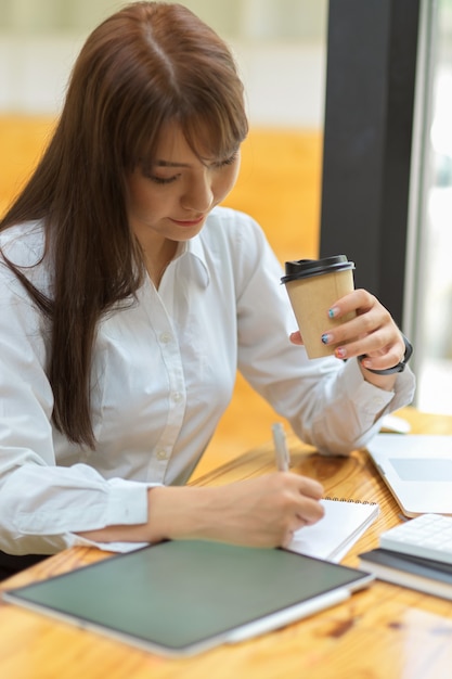 Secrétaire sérieuse et concentrée travaillant sur une tablette et buvant du café sur le lieu de travail