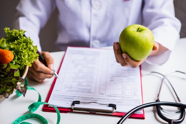 Section médiane du médecin tenant une pomme verte alors qu'il est assis avec un rapport et un stéthoscope sur la table