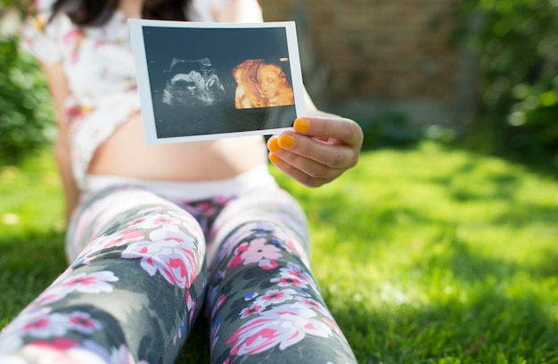 Photo section médiane d'une femme enceinte tenant une image à ultrasons contre l'abdomen assise sur l'herbe dans un parc