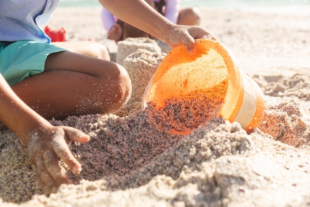 Section médiane d'un garçon biracial collectant du sable dans un seau avec sa sœur jouant à la plage par beau temps
