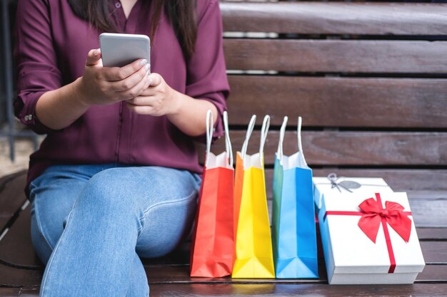 Photo section moyenne d'une femme adulte au milieu utilisant un téléphone portable tout en étant assise avec des sacs d'achat sur un banc