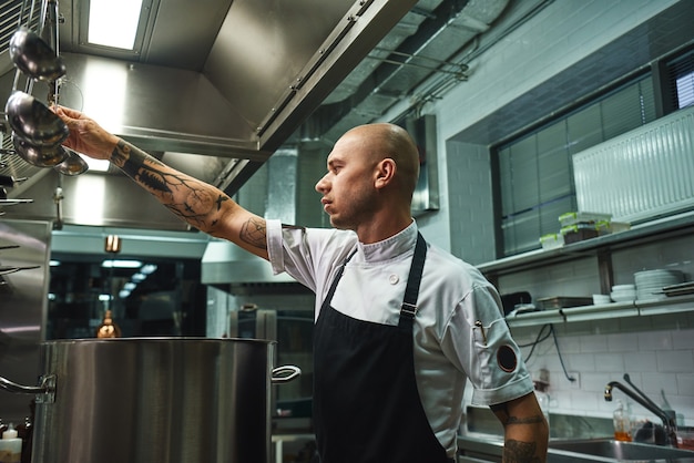 séduisant chef chauve en tablier prenant une louche pour la cuisson de la soupe en se tenant debout dans la cuisine d'un restaurant