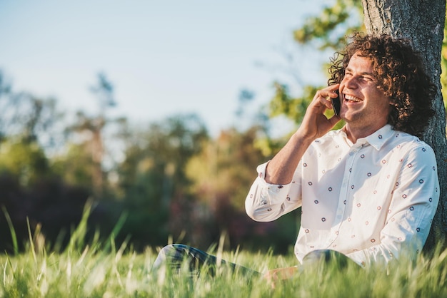 Séduisant homme heureux aux cheveux bouclés portant une chemise à la mode appelant sa petite amie pour lui dire qu'il l'attend dans le parc Espace de copie pour la publicité sur la technologie et la communication Mode de vie des gens