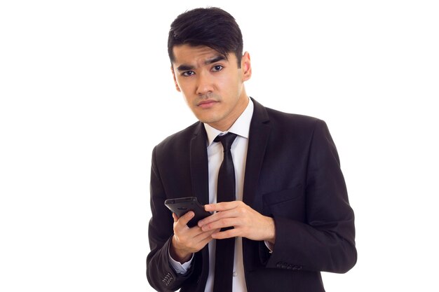 Séduisant jeune homme aux cheveux noirs en chemise blanche et costume noir avec cravate à l'aide de son téléphone