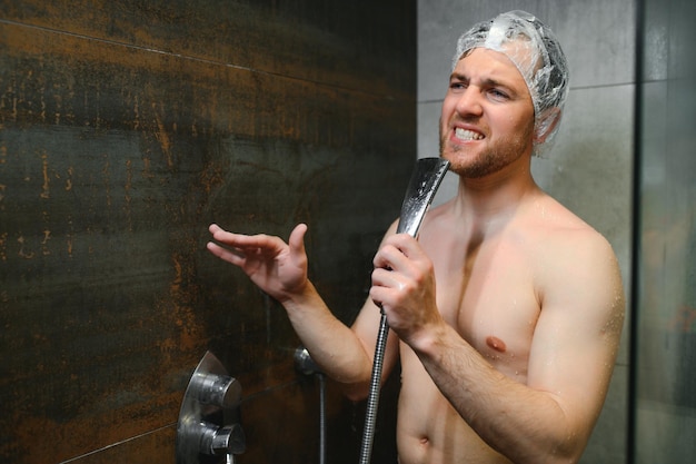 Photo séduisant jeune homme joyeux chantant tout en se lavant sous la douche, portant un bonnet de douche et tenant une pomme de douche