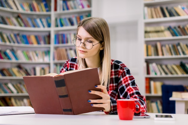 Séduisante blonde étudiante intelligente jeune fille à lunettes assis à la table, concentré tout en lisant un livre dans la bibliothèque de l'université. Étagère à livres avec différents livres sur l'espace