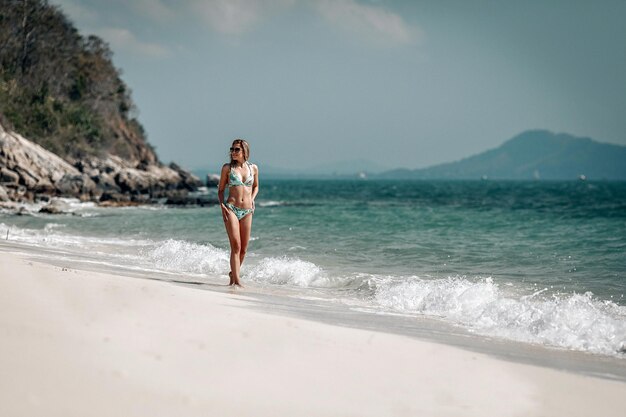 Séduisante blonde mince en bikini se promène le long de la plage de sable contre la baie bleue, journée ensoleillée, détente.