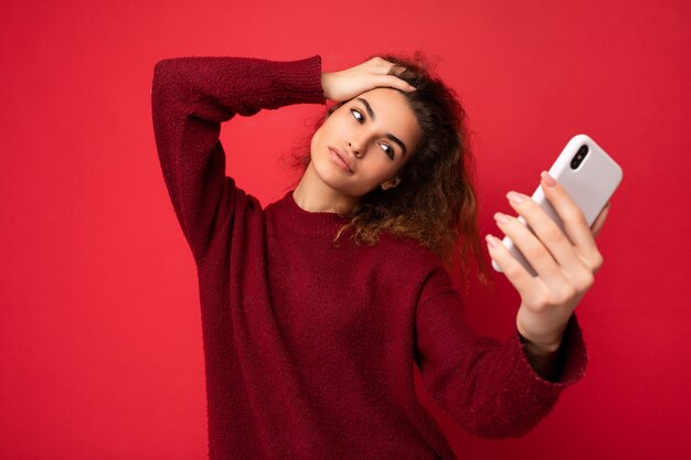 Séduisante charmante jeune femme souriante et heureuse tenant et utilisant un téléphone portable prenant un selfie portant des vêtements élégants isolés sur fond de mur