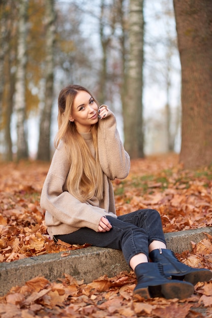 Séduisante élégante blonde souriante jeune femme portant un pull confortable, un jean noir à la mode et des bottes en cuir noir assis dans le parc de l'automne.