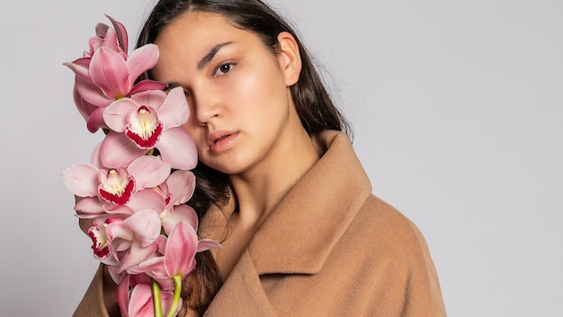 Séduisante femme blonde en veste marron qui pose en studio sur fond gris. modèle élégant en tenue de printemps décontractée pastel. Belle fille avec une peau saine et une orchidée de branche. Format panoramique 16:9.