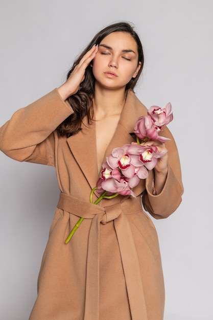 Séduisante femme blonde en veste marron qui pose en studio sur fond gris. Portrait de mode d'un modèle élégant en tenue de printemps décontractée pastel. Belle fille avec une peau saine et une orchidée de branche