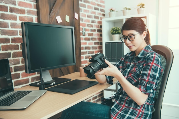 séduisante femme photographe élégante tenant une photo de travail d'examen de caméra d'affaires et choisissant une photo pour la retouche d'édition assis sur le bureau.