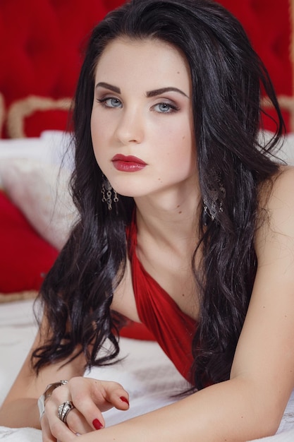 Séduisante femme en robe rouge se détendre sur un canapé dans un intérieur de luxe Modèle sexy dans une robe rouge Maquillage chic Brune avec de belles longues boucles