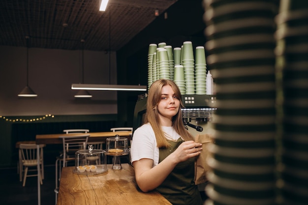 Séduisante jeune belle barista caucasienne en tablier souriant à la caméra au comptoir du café