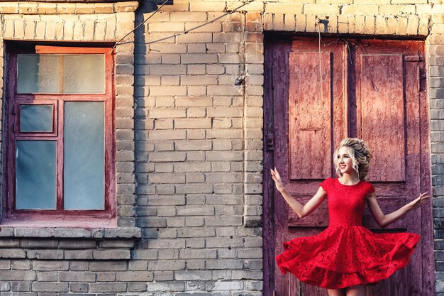 Séduisante jeune blonde en robe rouge posant sur le fond d'un vieux bâtiment en brique.