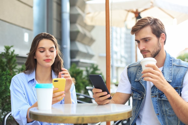 Séduisante jeune couple amoureux buvant du café assis à la table du café à l'extérieur, à l'aide d'un téléphone portable.