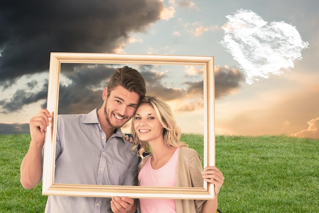 Séduisante jeune couple tenant un cadre photo contre le coeur des nuages