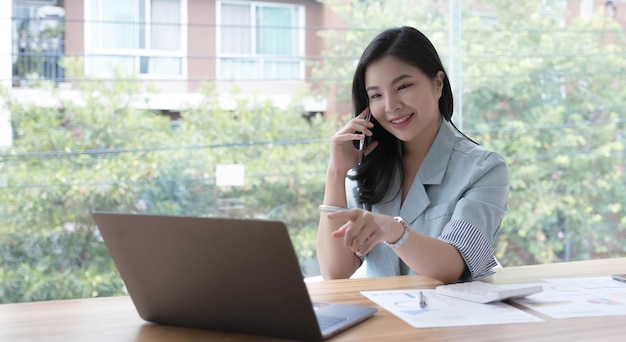 Séduisante jeune femme d'affaires asiatique souriante se relaxant au bureau travaillant sur un ordinateur portable parlant sur un téléphone mobile