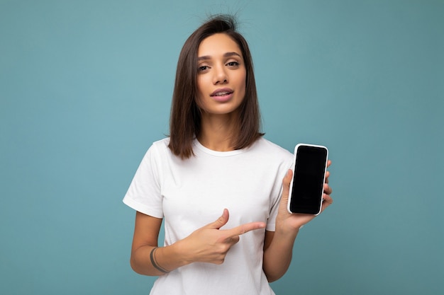 Séduisante jeune femme brune bonne recherche portant un t-shirt blanc debout isolé sur bleu