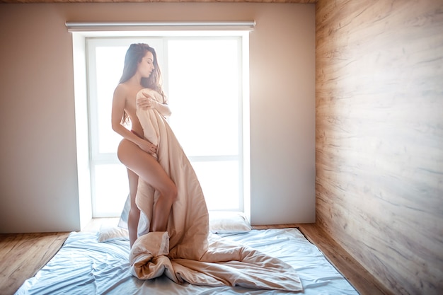 Photo séduisante jeune femme brune nue sur le lit le matin. corps de couverture de beau modèle sexy avec obturé. debout sur le lit. seul dans la chambre