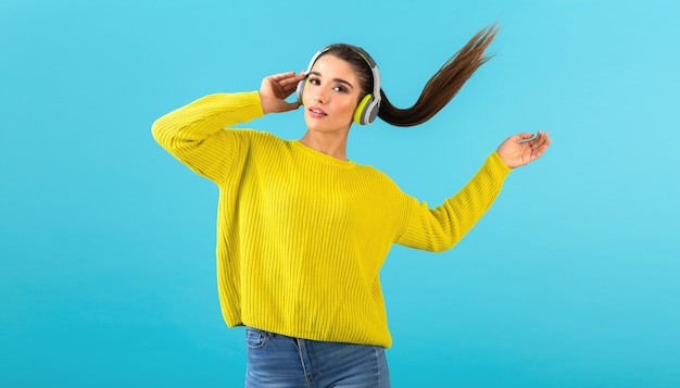 Séduisante jeune femme élégante écoutant de la musique dans des écouteurs sans fil heureux portant un chandail tricoté jaune mode de style coloré posant isolé sur fond bleu en agitant la queue de cheveux longs