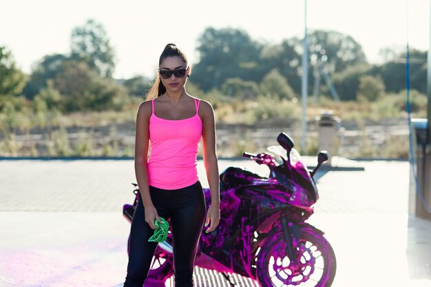 Séduisante jeune femme en tshirt rose pose près de moto de sport au lave-auto en libre-service dans le