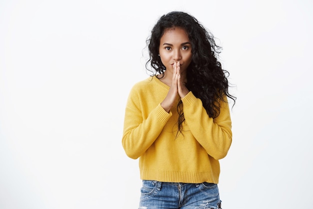 Séduisante jeune fille aux cheveux bouclés afro-américaine en chandail d'automne jaune presse les paumes ensemble dans la prière près des lèvres regarde caméra sincère mendicité espoir ami aide suppliant fond blanc