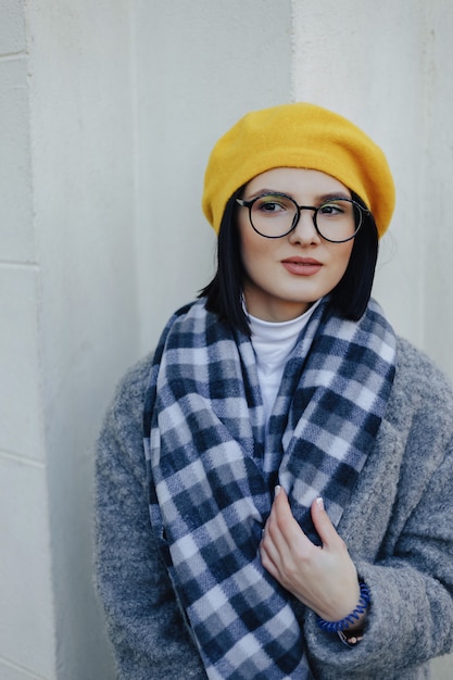 Séduisante jeune fille à lunettes en manteau et béret jaune sur un fond clair simple