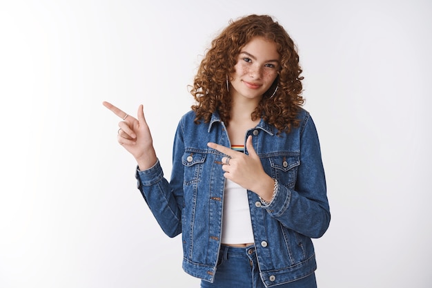 Séduisante jeune fille rousse aux cheveux bouclés, boutons de rousseur souriant largement invitant un ami café végétalien pointant l'index gauche demandant un essai souriant joyeusement fond blanc