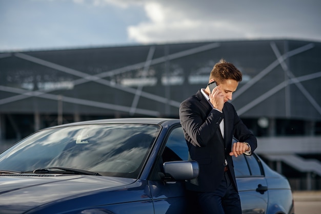 Séduisante jeune homme d'affaires prospère dans un costume d'affaires et regarder à la main parler sur smartphone près de sa voiture