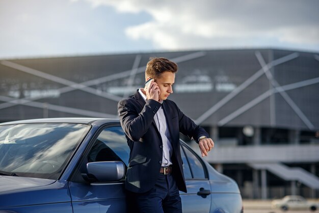 Séduisante jeune homme d'affaires prospère dans un costume d'affaires et regarder à la main parler sur smartphone près de sa voiture