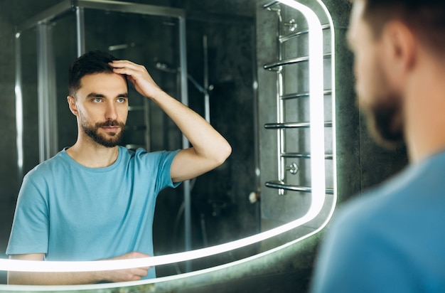 Séduisante jeune homme aux yeux bleus touchant ses cheveux devant le miroir le matin