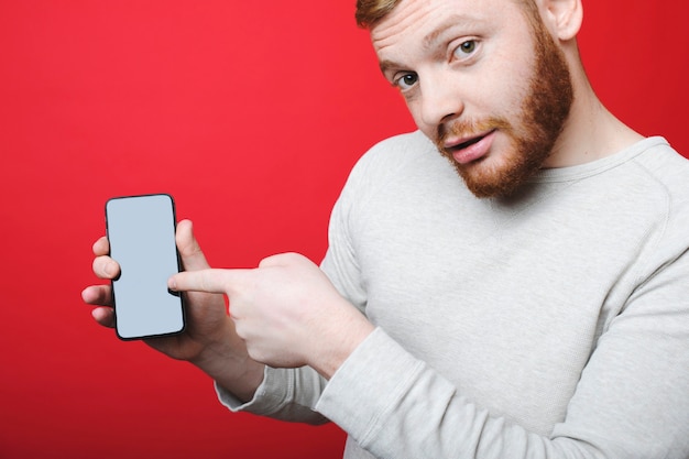 Séduisante jeune homme avec barbe au gingembre regardant la caméra et pointant sur smartphone moderne