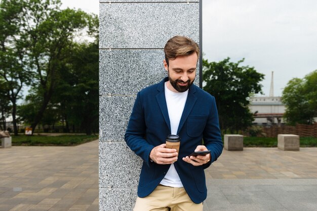 Séduisante jeune homme barbu souriant portant une veste à l'aide d'un téléphone portable tout en se tenant à l'extérieur de la ville et en buvant du café à emporter