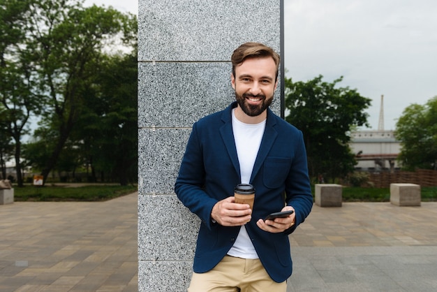 Séduisante jeune homme barbu souriant portant une veste à l'aide d'un téléphone portable tout en se tenant à l'extérieur de la ville et en buvant du café à emporter