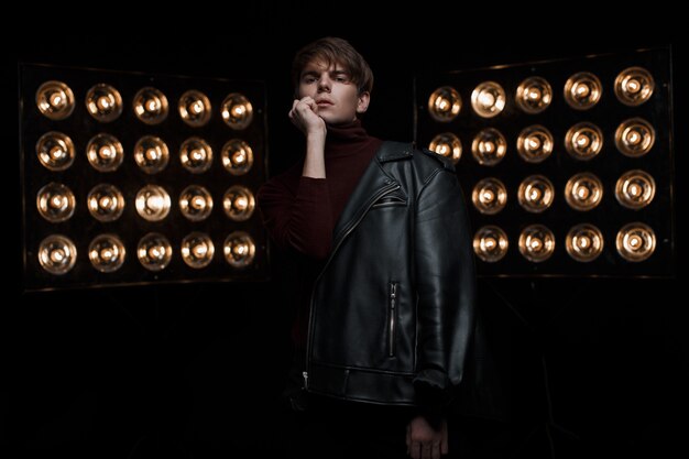 Séduisante jeune homme dans une veste en cuir à la mode noire dans un golf élégant rouge et un jean noir se dresse et pose dans une pièce sombre près de lampes professionnelles électriques vintage lumineuses. Modèle de type charmant