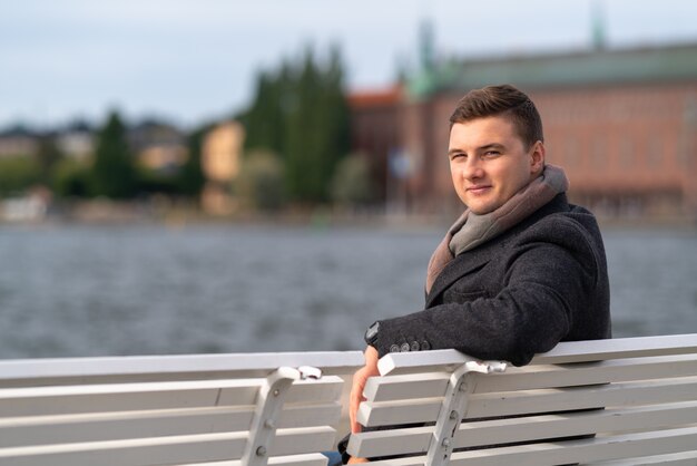 Séduisante jeune homme en manteau chaud et écharpe assis sur un banc à l'extérieur donnant sur l'eau agitée se tournant pour sourire à la caméra