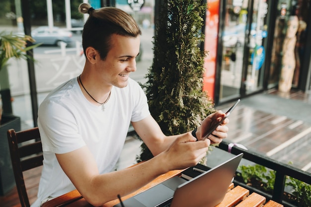 Séduisante jeune homme souriant et parcourant la tablette moderne alors qu'il était assis à table dans un café en plein air près d'un ordinateur portable