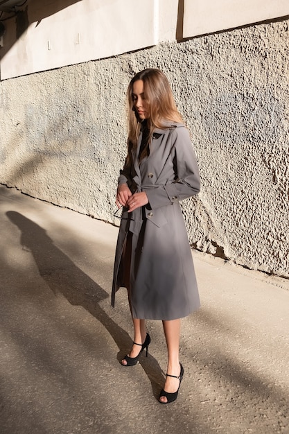 Séduisante jeune mannequin asiatique en trench-coat gris debout près du mur à l'extérieur