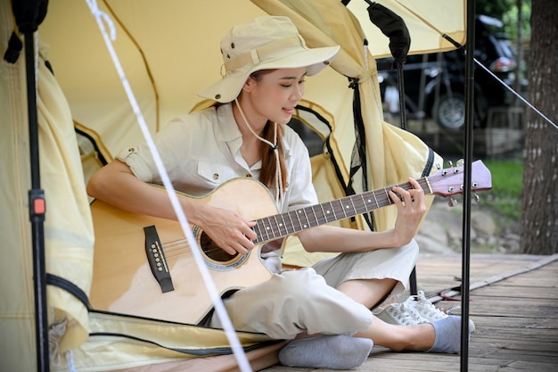 Séduisante jeune voyageuse asiatique assise devant sa tente de camping et jouant de la guitare