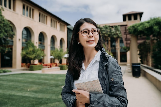 Séduisante souriante charmante étudiante asiatique jeune fille avec livre scolaire relaxant marchant à l'extérieur de l'université. college lady profitez du soleil dans le parc de stanford entouré d'herbe verte et d'arbres.