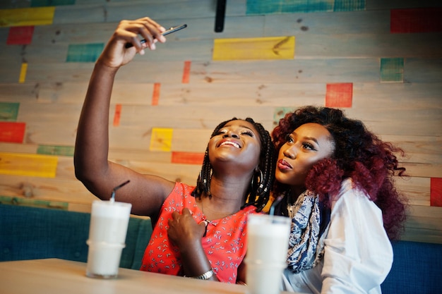 Séduisantes amies afro-américaines de deux filles assises au café avec un café au lait et faisant un selfie sur un téléphone portable