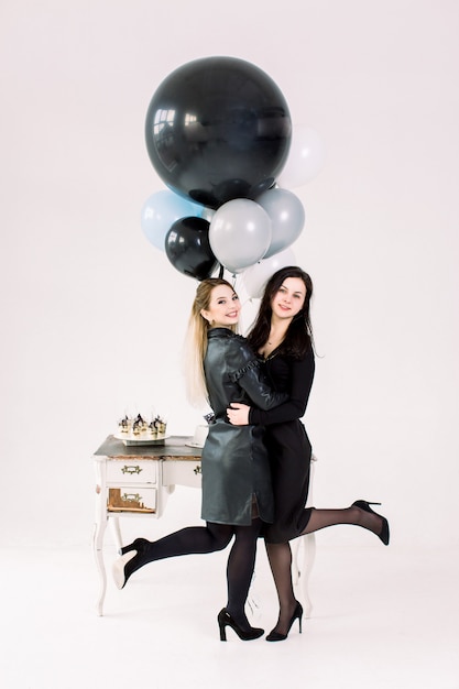 Séduisantes jeunes filles en robes noires posant devant une table vintage blanche avec gâteau et martini, tenant des ballons à air big coloré, sur fond blanc. Bonne année, fête d'anniversaire