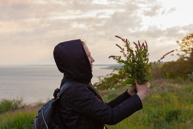 Séjour fille en imperméable avec sac à dos bouquet de fleurs sauvages sur fond de paysage marin nature Tendance hyper voyage local