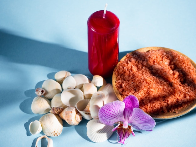 Sel de bain orange dans une soucoupe avec coquillages, bougie rouge et fleur sur fond bleu avec une ombre d'une plante tropicale. Copyspace. Spa, détendu, été