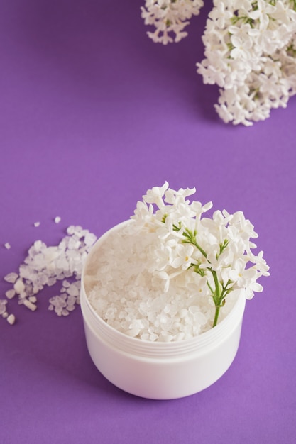 Sel de mer blanc dans un pot en plastique et fleurs lilas blanches sur fond lilas concept cosmétique de soins corporels naturels