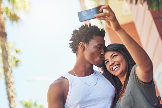 Selfie baiser et couple interracial avec téléphone dans la ville date et bonheur avec mémoire ensemble amour de la technologie 5g et homme et femme noirs avec affection pour une photo mobile avec un sourire