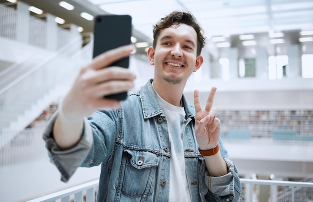 Selfie de bibliothèque et homme d'université avec signe de paix pour la mise à jour du mode de vie des médias sociaux ou un article de blog avec une éducation heureuse Étude de recherche et étudiant en doctorat ou personne avec signe emoji pour photo de profil